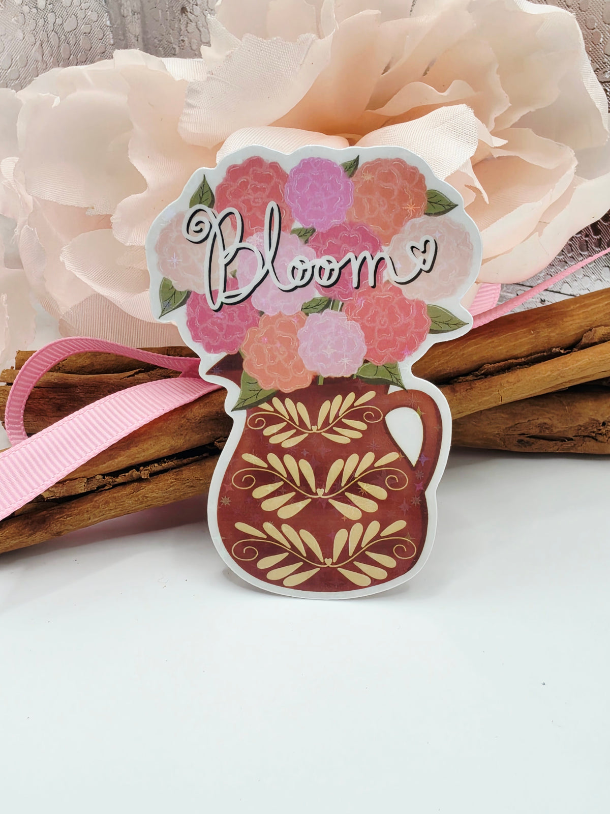 A decorative die-cut ornament shaped like a flower and a clay vase, filled with pink flowers, green leaves, and the word 'Bloom' in script, hanging beside cinnamon sticks and a pale pink ribbon.