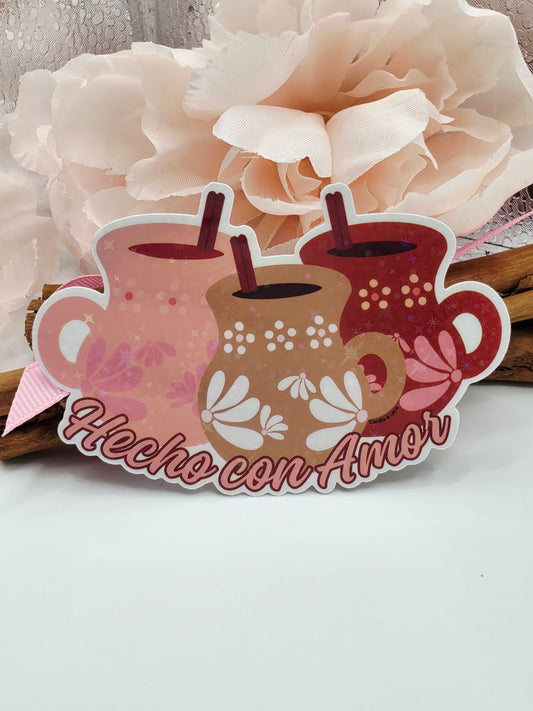 A decorative die-cut sign shaped like 3 style mugs lined up together with Spanish text reading 'Hecha con Amor,' shown next to a large cream paper flower against a plain light background.