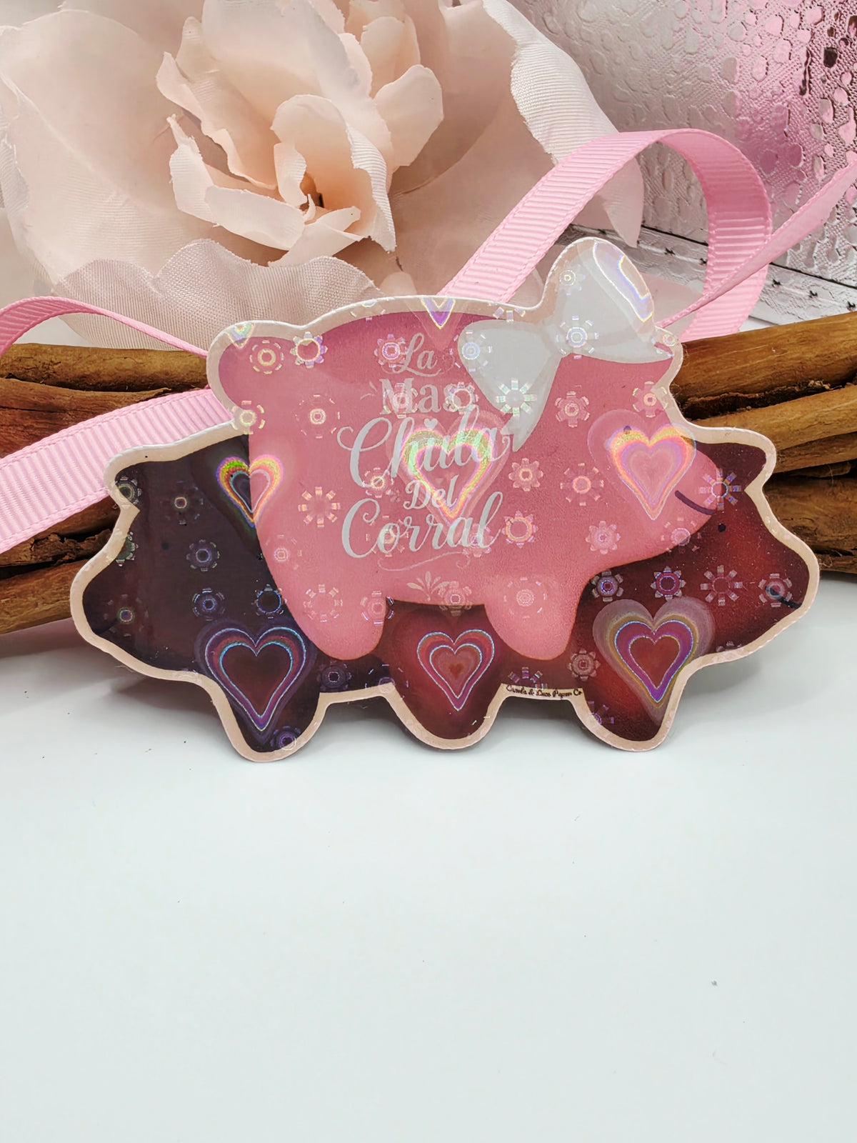 3 different colored pigs inspired by marranitos, a type of mexican sweet bread. The pigs are dark brown, light brown and pink. The pink pig wears a white and pink bow. The caption on the sticker reads, "La mas chula del corral." Vynl Sticker is laminated with a heart Holographic design. 