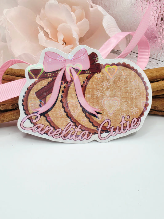 A decorative die-cut sticker or label featuring a layered pastry illustration with the words 'Canelita Cutie' in pink script, attached to a pink ribbon and displayed among pale flowers and wooden sticks on a white background.