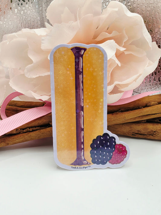 A novelty sticker shaped like a sandwich cookie, featuring two golden-yellow layers, a purple filling, and blackberries on top, styled with a large cream flower, pink ribbon, and cinnamon sticks on a white background.
