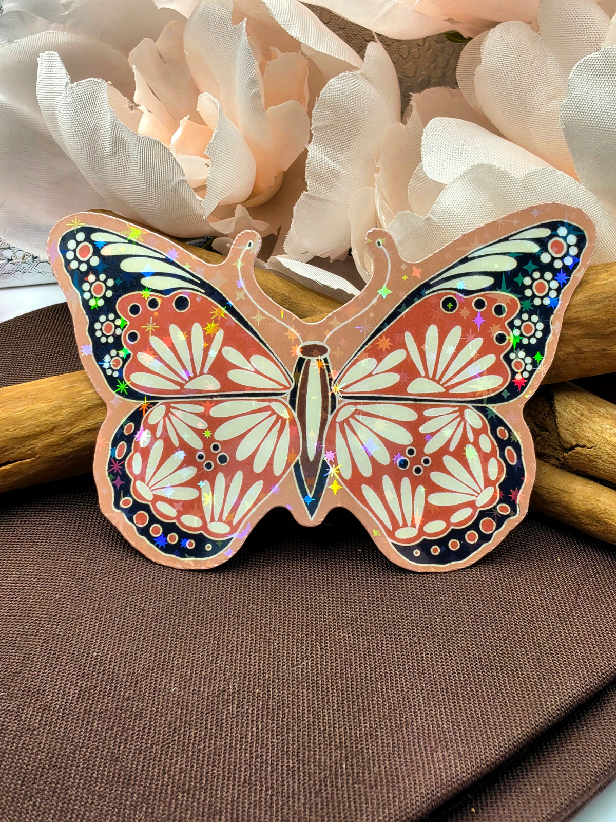 A butterfly-shaped decorative item with a holographic foil finish, featuring orange, white, and black patterns, displayed on a brown textured surface with white flowers in the background.